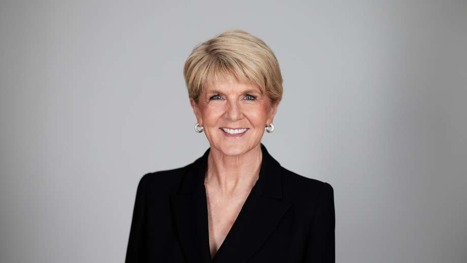 A woman standing in front of a grey background with short hair and a black blazer smiles.