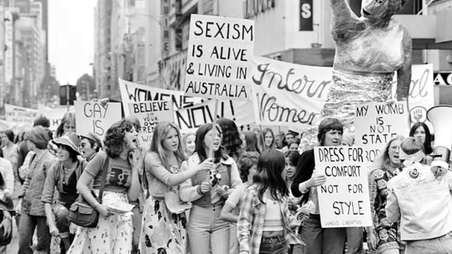 Black and white photograph from 1975 shows large group of women marching through Melbourne and holding banners with messages in support of women's rights on them.