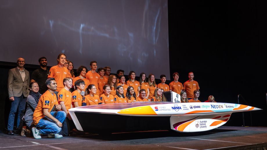 Photo of the solar racing team - all wearing orange shirts - standing behind their new car