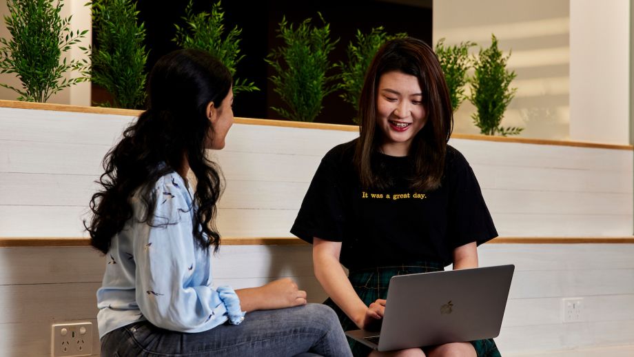 Two women smiling, talking and working on their laptops.