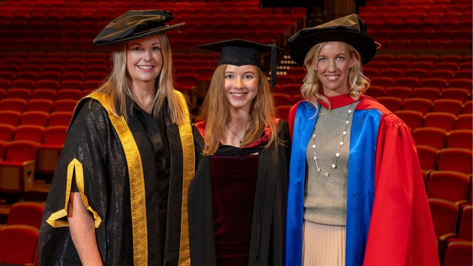 Three women at a graduation ceremony pose for a picture in front of an auditorium.