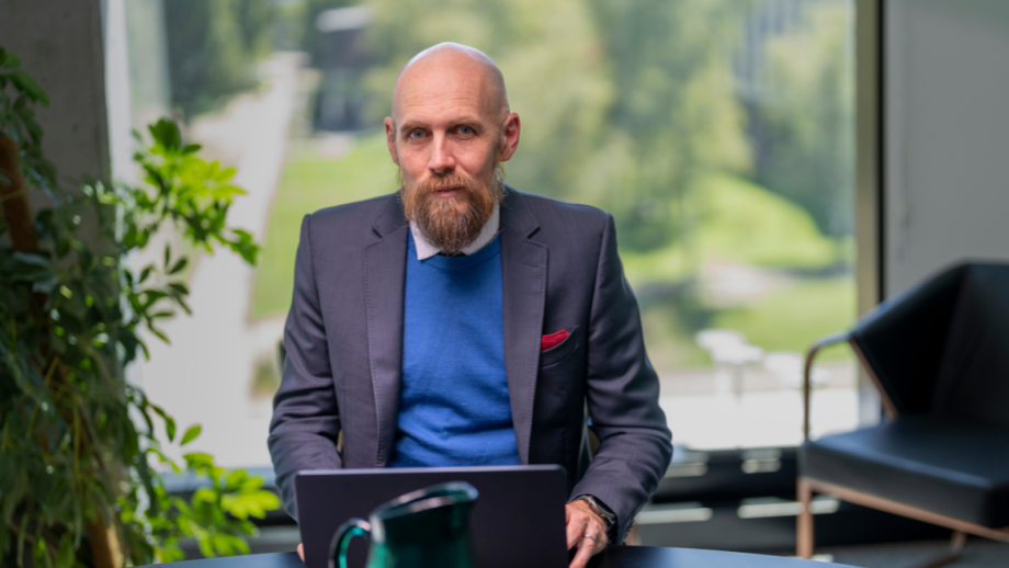 Professor Nicholas Biddle seated at a desk with a laptop, in front of a window, looks at the camera.