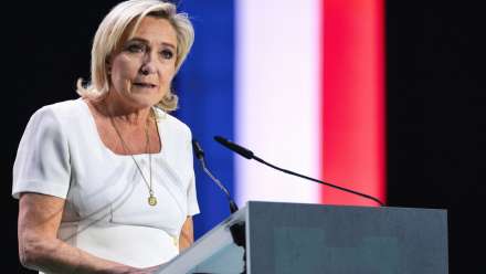 Marine Le Pen, leader of the far-right National Rally party in France. She is standing behind a podium and there is a French flag in the background.