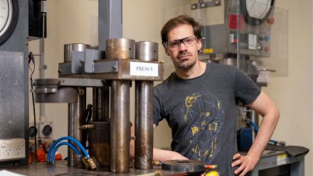 Dr Michael Anenburg is wearing safety goggles and is leaning against a desk next to lab equipment in the Research School of Earth Sciences