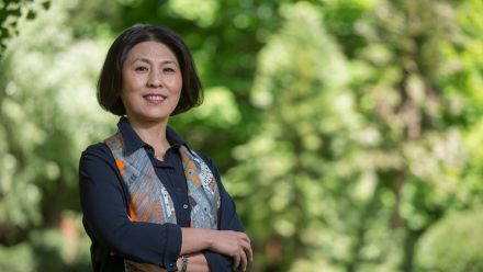 Distinguished Professor Xuemei Bai has her arms crossed and is smiling. There are trees behind her