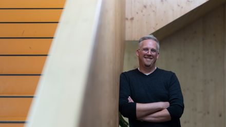 Associate Professor Mathew Davies standing in a hall, smiling at the camera, leaning against a wall with his arms folded.