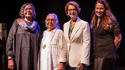 Four women smiling and posing together on a stage at a formal event.