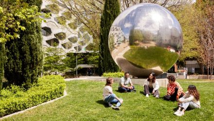 a group of student sits on a lawn in front of a reflective ball sculpture. 