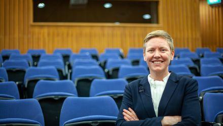 Heidi Yates smiles at the camera with her arms crossed in a lecture theatre with rows of blue seats.