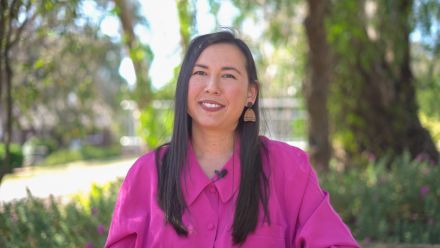 Sophia Hamblin-Wang smiles at the camera, wearing a bright pink shirt, with trees and greenery in the background.