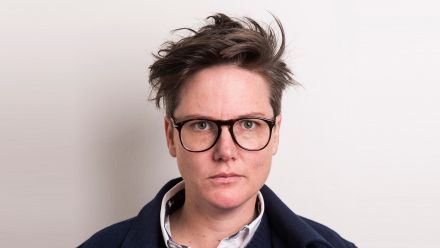 Hannah Gadsby looks directly at the camera, wearing black-framed glasses and a navy jacket against a plain white background.