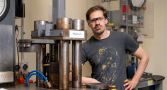 Dr Michael Anenburg is wearing safety goggles and is leaning against a desk next to lab equipment in the ANU Research School of Earth Sciences