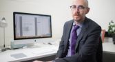 Professor Nick Biddle sits at his desk wearing a purple shirt and tie with a blazer. There is a computer behind him.