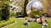 a group of student sits on a lawn in front of a reflective ball sculpture. 