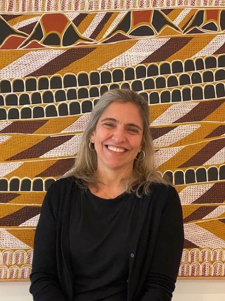 A smiling person with light shoulder length hair standing in front of an artwork.