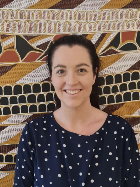 A woman with dark brown hair tied up in smiling in front of artwork.