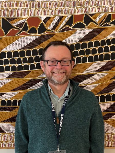 A man with a green jumper, glasses and beard smiling in front of indigenous artwork