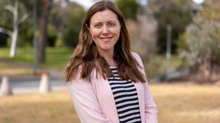 Dr Claire Hansen is wearing a pink blazer and a black and white striped shirt underneath. She is standing outside in front of trees