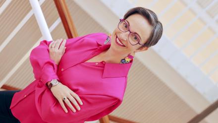 A woman with short hair wearing a pink blouse and glasses stands with her arms folded. 