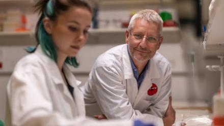 Photo of Alex leaning on a bench in a lab, talking to a student. 