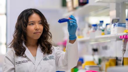 Dr Chelisa Cardinez inspects a test tube containing an unknown blue substance. She is wearing a white lab coat.