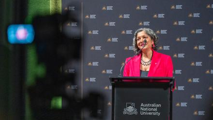 A woman with short black hair wearing a pink blazer speaks at a lectern.