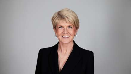 A woman standing in front of a grey background with short hair and a black blazer smiles.