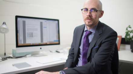 Nick sits at a desk facing the camera. He is wearing a suit and a purple tie.
