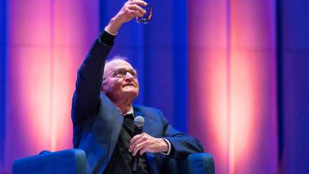 Photo of Professor Brian Walker saluting Will by raising his glass. He's wearing a suit and holding a microphone.