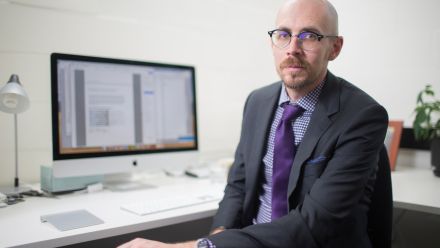 Professor Nick Biddle sits at his desk wearing a purple shirt and tie with a blazer. There is a computer behind him.