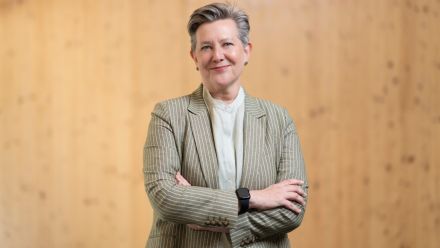 Professor Joan Leach stands with her arms crossed, smiling. She is wearing a gray blazer.