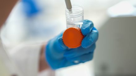 Close up of person in gloves and lab coat holding a sample container and syringe. 