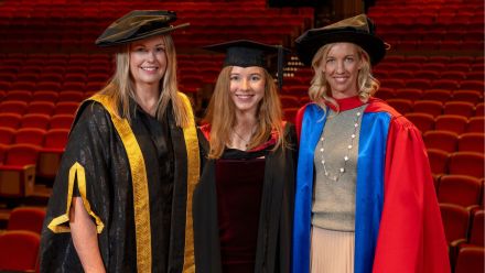 Three women at a graduation ceremony pose for a picture in front of an auditorium. 
